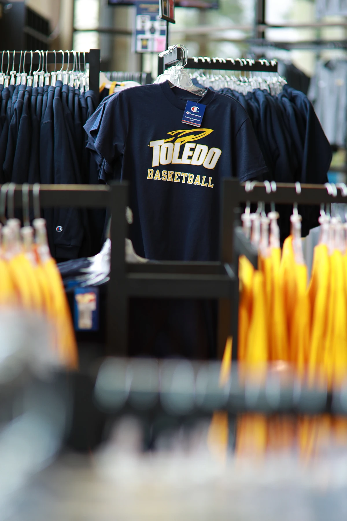 A navy blue Toledo Basketball t-shirt hangs prominently in a store display with other clothing on racks nearby.
