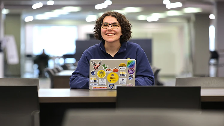 A person with curly hair and glasses smiles while sitting at a table with a sticker-covered laptop in a library or study area.