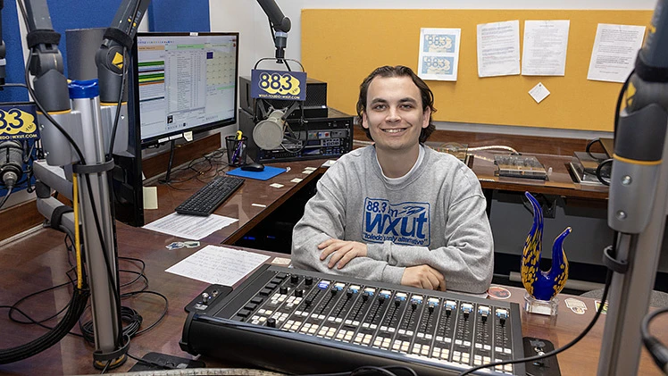 A person is sitting at a radio station control panel, smiling at the camera, with equipment and monitors surrounding them.