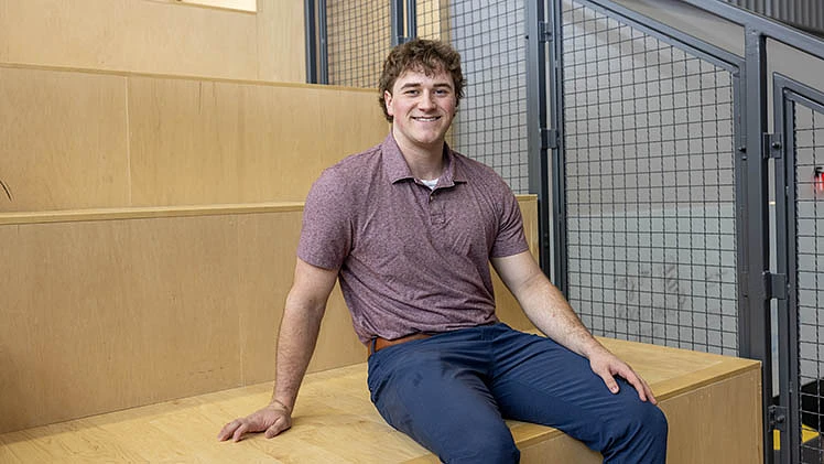 A person in a maroon shirt and blue pants is sitting on wooden steps, smiling at the camera.
